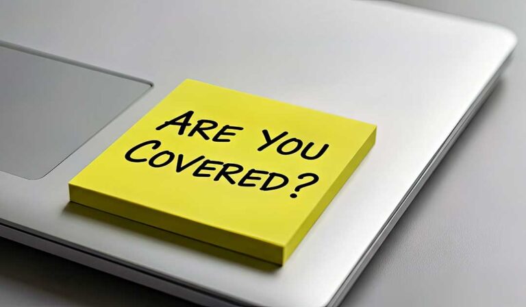 A person reviews insurance paperwork and takes notes at a kitchen table with a laptop and printed documents.